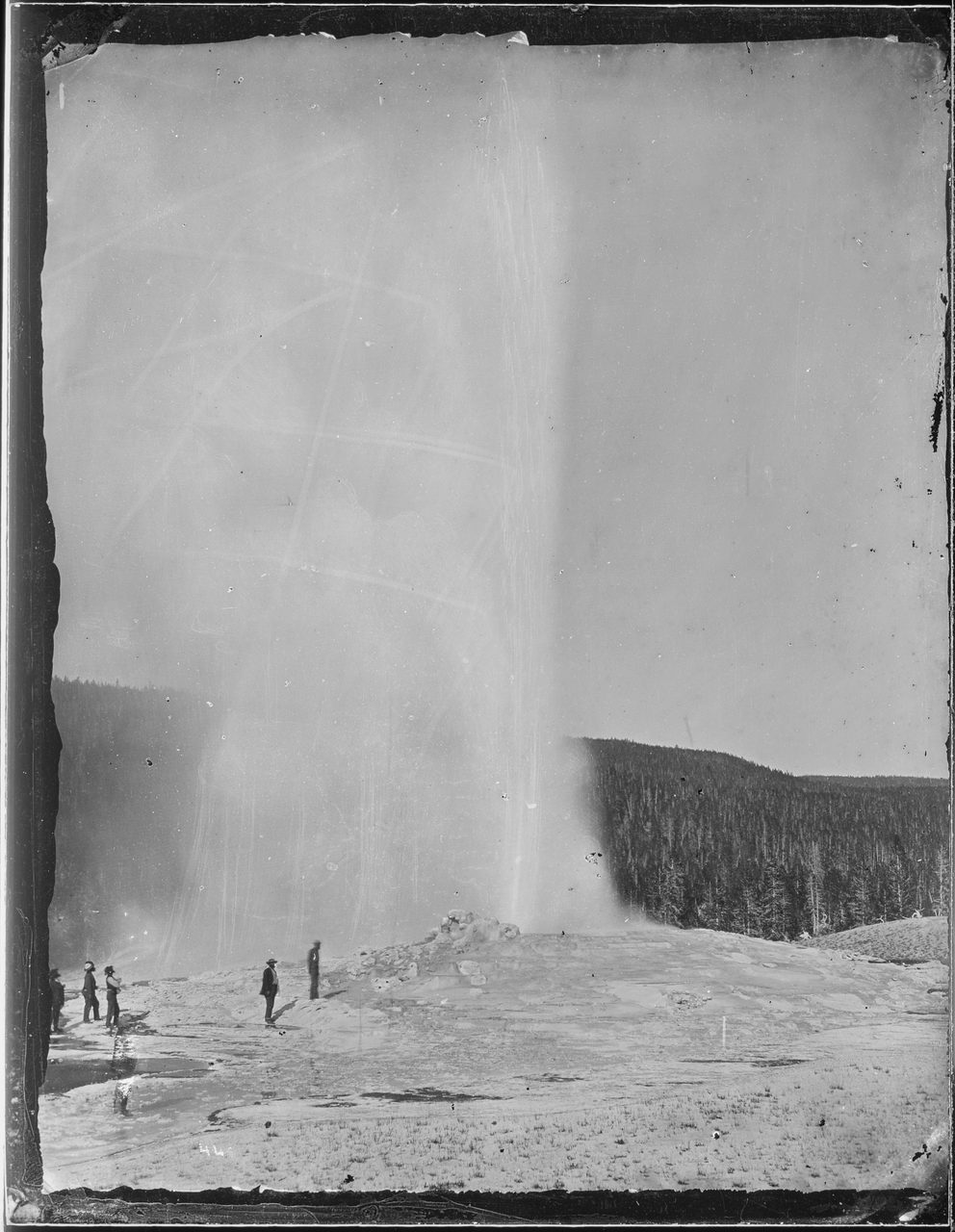 A damaged 1872 glass-plate photograph of Old Faithful erupting, a tall pale column of water and steam, with several figures in period dress standing on the sinter nearby