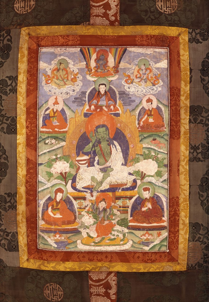 A thangka in silk brocade borders showing a green skinned yogi seated on a white animal skin, his right hand cupped at his ear, a single white cloth draped across his body, surrounded by smaller lineage figures